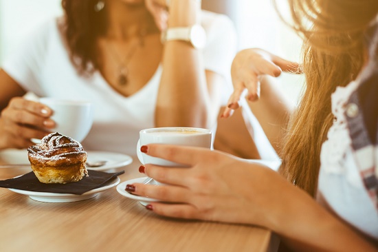 Shown: Two women seated at a table drinking coffee and talking. 