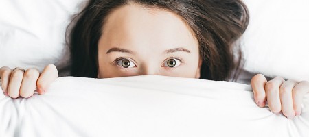 Woman laying in bed, hiding the bottom of her face with a white bedsheet.