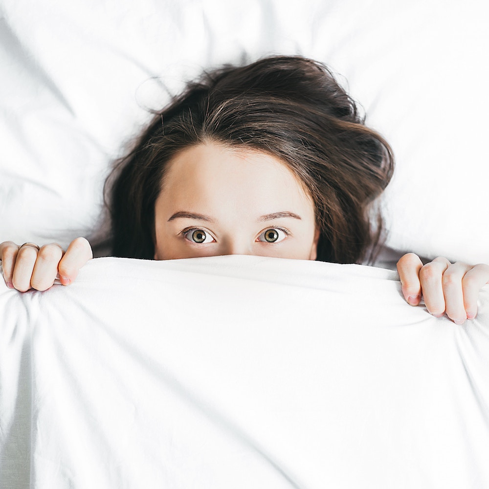 Woman laying in bed, hiding the bottom of her face with a white bedsheet.