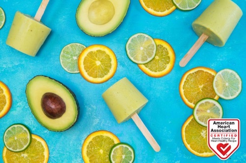 Shown on a table, an avocado cut in two, an avocado and banana frozen popsicle, and several orange and lime slices. 