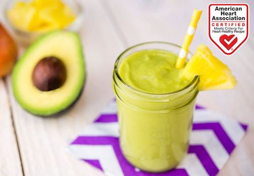 Shown on a table, half an avocado with the pit in the center. a plate with crushed mango, ,and a avocado and mango smoothie in a mason jar with a straw and a lemon slice on the rim.