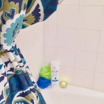 Shown: Multi-colored shower curtain opened to show two bath sponges in blue and green, an Ivory Body wash, and a lit tea light, on tub shelf.