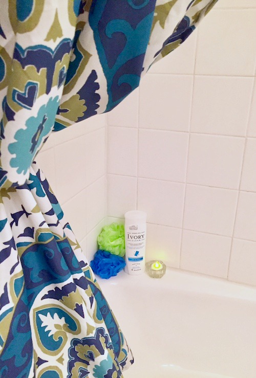 Shown: Multi-colored shower curtain opened to show two bath sponges in blue and green, an Ivory Body wash, and a lit tea light, on tub shelf.