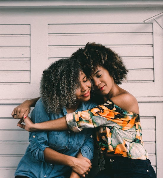 Shown: Two women of color seated and hugging. | PC: Hian Oliveira on UnSplash