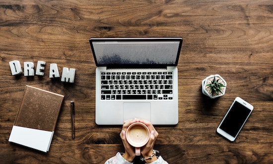 On a wooden table, two hands holding a coffee mug, a laptop, iPhone, pen, small plant and the word D R E A M. Photo Credit: rawpixel on UnSplash.