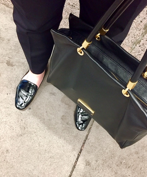 Shown: Lower half of woman standing on sidewalk. Features her black patent leather Dolce Vita Flats and Marc by Marc Jacobs handbag.