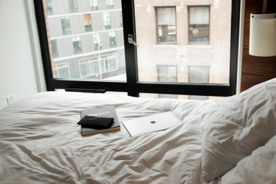 Arlo Soho hotel room - an Apple Macbook and a Sketch drawing book on top of a queen bed are shown in front of a window. 